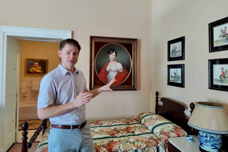A man looks forward at the camera while gesturing toward a painting of a woman from the 19th century