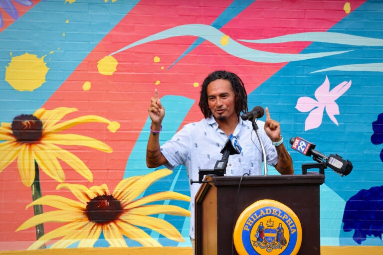 A man stands at a podium in front of a mural on a wall featuring yellow flowers and varying colors