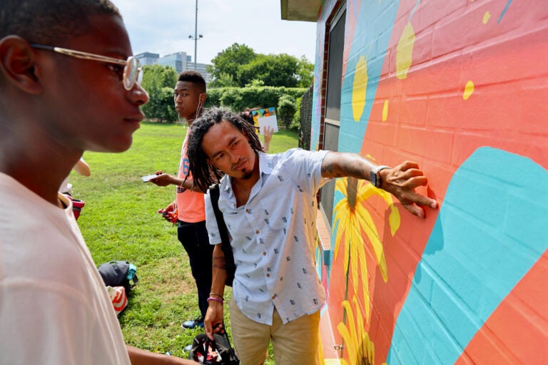 A man puts his hand on the wall with a colorful mural while another man looks at the mural