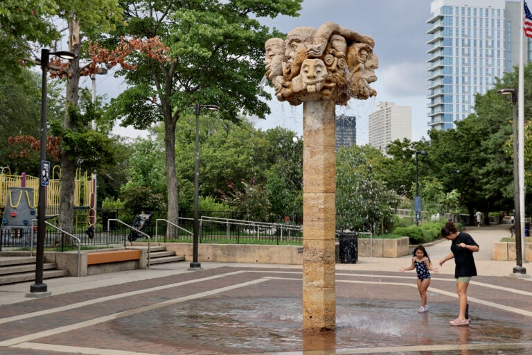 A tall sculpture with several heads on top cascades water down on a child and woman underneath it