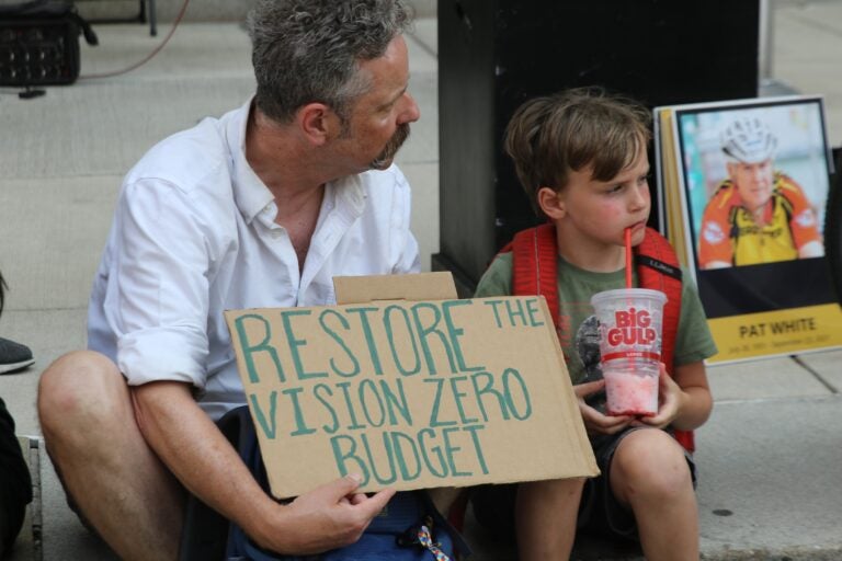 a man holds a sign reading RESTORE THE VISION ZERO BUDGET next to a child