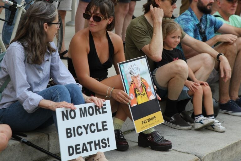 people sit, two holding signs, one reading NO MORE BICYCLE DEATHS and the other being a memorial for Pat White