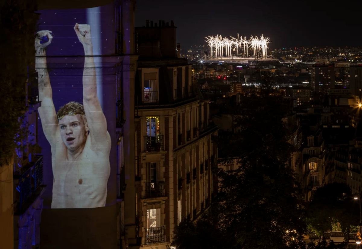 A photograph of swimming gold medalist Leon Marchand of France celebrating winning the men's 200 meter individual medley final is projected in Montmartre overlooking the fireworks of the closing ceremony