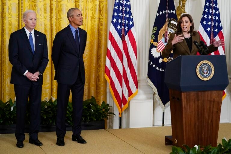 Kamala Harris speaking at a podium with Joe Biden and Barack Obama standing beside her