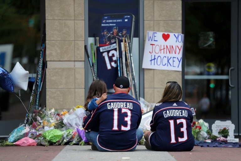 Two people wearing Gaudreau jerseys sit and hold a kid at the memorial