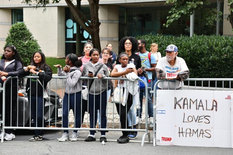 A lone trump supporter with a sign reading KAMALA LOVES HAMAS stands behind Harris fans behind a fence