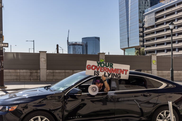 a person driving shouts out of a megaphone and holds a sign reading YOUR GOVERNMENT IS LYING