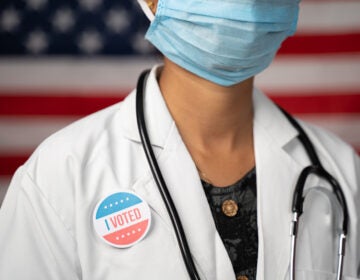 Doctor or nurse medical mask and stethoscope wearing placing I voted sticker to her apron - concept of US election with US flag as background