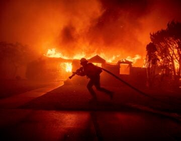 A firefighter dragging a fire hose in front of a house completely engulfed in flames