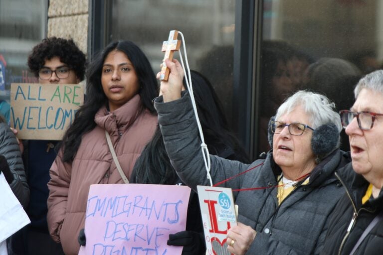 Protesters hold a cross and signs reading ALL ARE WELCOME and IMMIGRANTS DESERVE DIGNITY