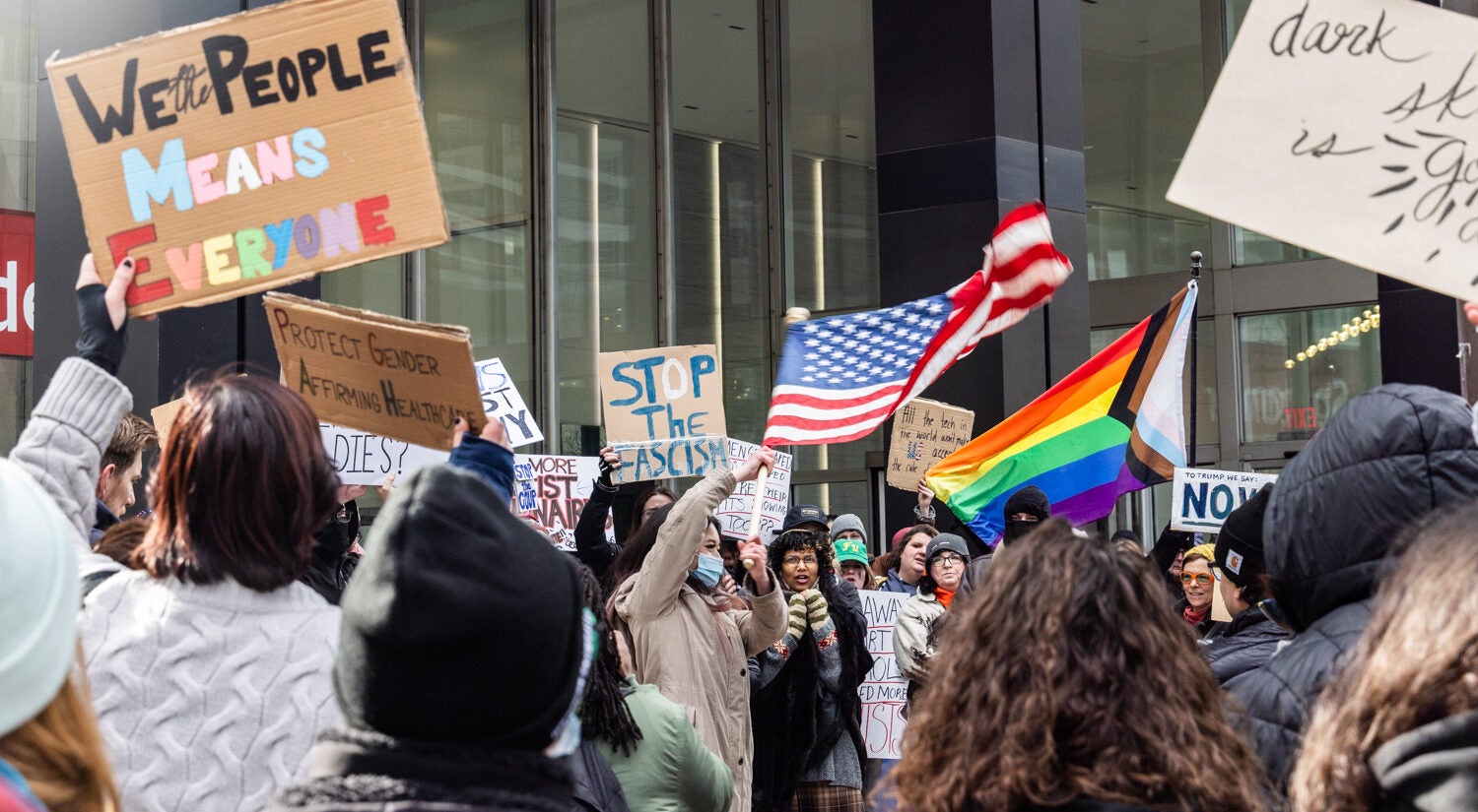 a protester waves an American flag, and a sign reads WE THE PEOPLE MEANS EVERYONE