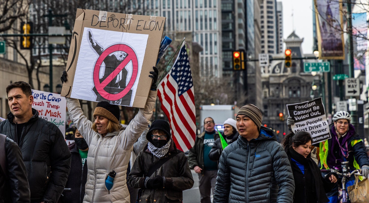 protesters walking in the street. a sign reads DEPORT ELON and shows his Nazi-like salute