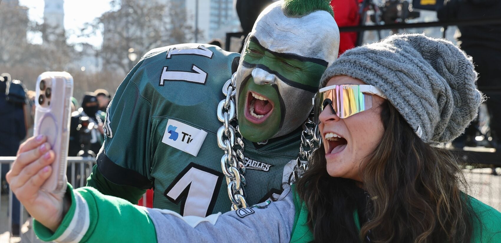 A woman takes a selfie posing with a guy in full face-paint and mohawk and eagles jersey