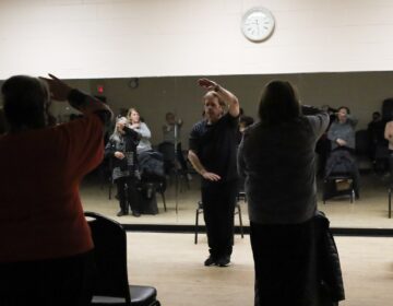 A man in the center of a large room does tai chi, and several people are doing it with him