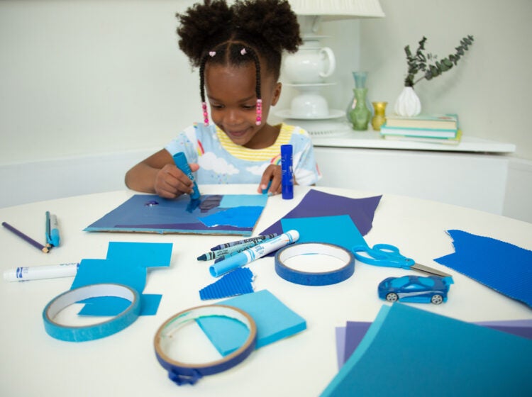 A child sitting at a table doing arts and crafts with blue paper, blue tape, and blue markers.