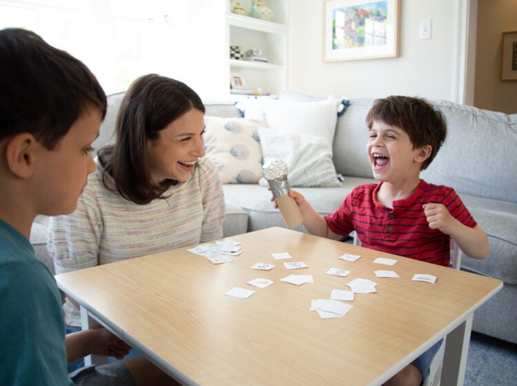 A child excited and happy sitting at a table with another child and an adult. The child is holding a microphone made of tinfoil and a toilet paper role. There are paper cars on the table.