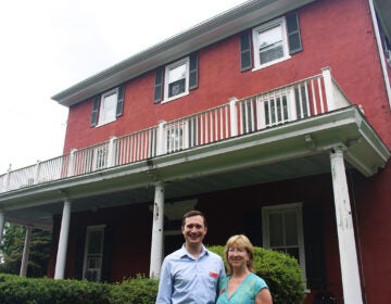 A man and a woman smile at the camera outside a red house