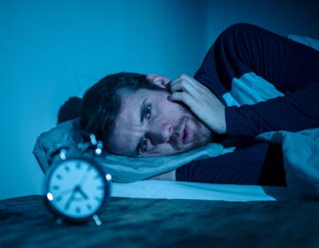 Sleepless and desperate young caucasian man awake at night not able to sleep, feeling frustrated and worried looking at clock suffering from insomnia in stress and sleeping disorder concept.