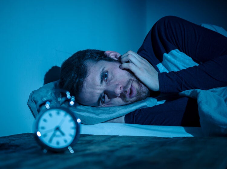 Sleepless and desperate young caucasian man awake at night not able to sleep, feeling frustrated and worried looking at clock suffering from insomnia in stress and sleeping disorder concept.
