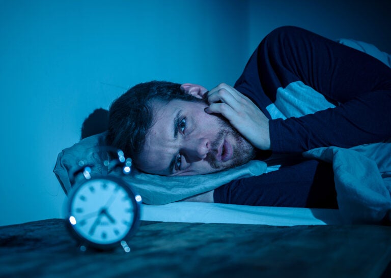 Young Man In Bed Staring At Alarm Clock Trying To Sleep Feeling Sleepless and desperate young caucasian man awake at night not able to sleep, feeling frustrated and worried looking at clock suffering from insomnia in stress and sleeping disorder concept.