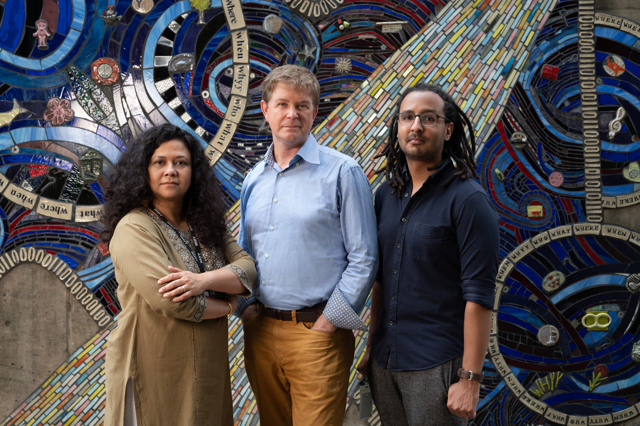 Three people from the WHYY News staff posing in front of a mosaic mural