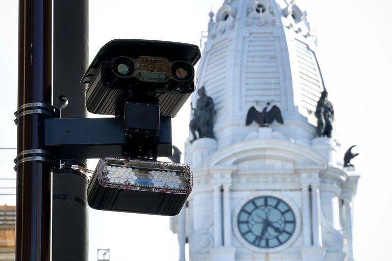Close-up shot of a speed camera attached to a light pole, with City Hall in the background