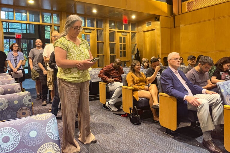 A woman speaks at a podium during the meeting, with other people lining up behind her for their turn.