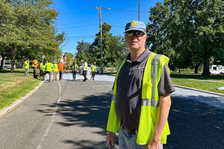 Steve Lorenz posing for a photo on the street, while workers lay down the coating behind him