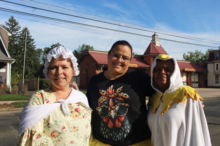 Three women, one on the left dressed in 18th century grab, one dressed in a T-shirt in the middle, and one on the right dressed as a chicken, smile for the camera