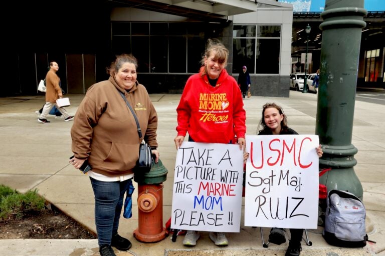 Three people with two signs, one reading TAKE A PICTURE WITH THIS MARINE MOM PLEASE!! and the other reading USMC SGT MAJ. RUIZ