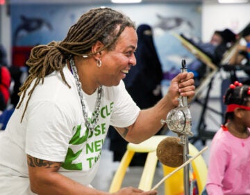 Dendê Macêdo plays a percussion instrument made from a broom, a bell, a coconut and a spring alongside a child