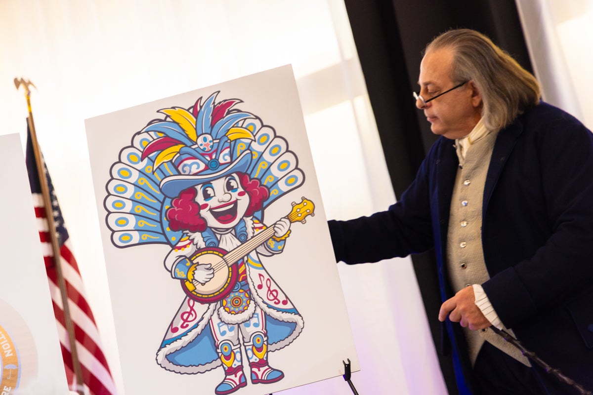 A Ben Franklin historical reenactor shows a drawing of a doll with red hair and colorful dress to be the Mummers 2026 plushie
