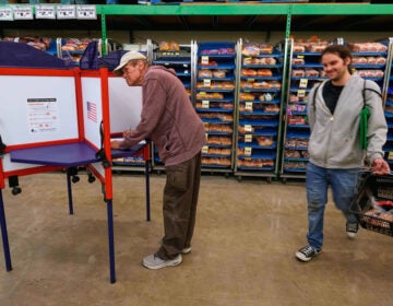 A person votes at a grocery store while another person walks by with a basket of food items