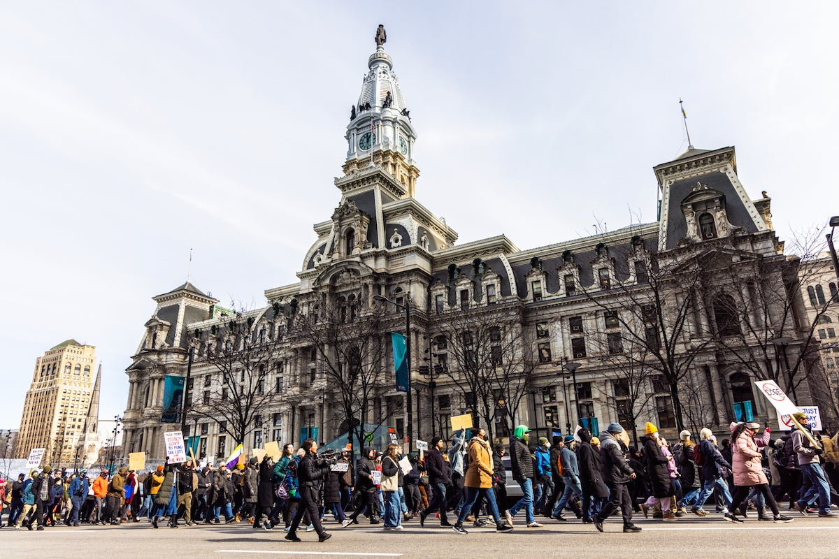 A wide photo of a long line of people, dressed in winter coats, marching around Philadelphia City Hall, which can be seen in the background. Many are holding handmade signs with slogans against President Trump.