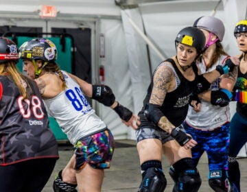04 11 2025 k paynter roller derby stills-8.jpg Five women from the Brandywine Roller Derby team, wearing athletic clothes, kneepads, elbow pads, and wrist guards are in the middle of practicing for roller derby. They are also wearing helmets, which have a variety of colorful stickers on them.