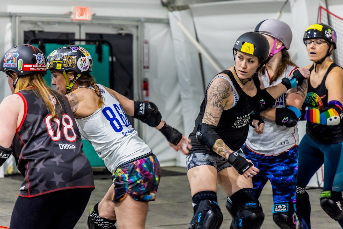 Five women from the Brandywine Roller Derby team, wearing athletic clothes, kneepads, elbow pads, and wrist guards are in the middle of practicing for roller derby. They are also wearing helmets, which have a variety of colorful stickers on them.