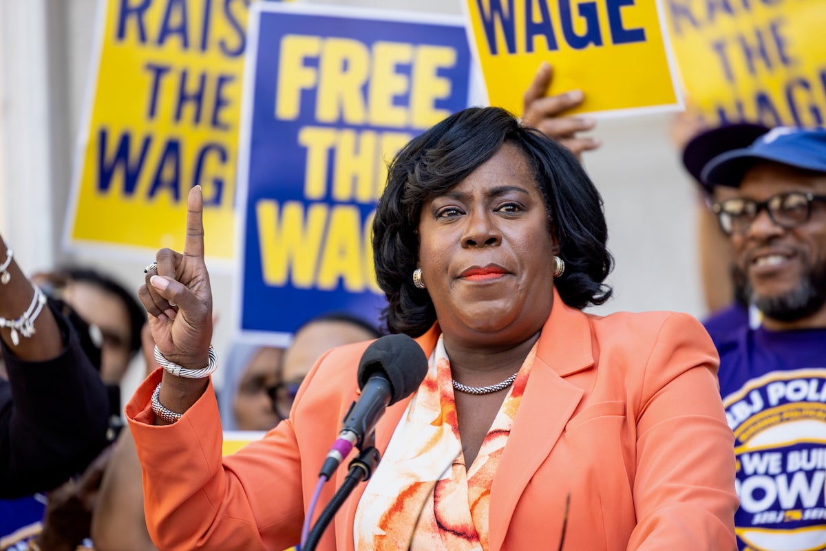 Philadelphia Mayor Cherelle Parker is holding up one finger, as she pauses for a moment while speaking into a microphone at a rally for 32BJ SEIU workers. Some workers stand behind her wearing blue shirts with the union's logo, and holding signs that say 'RAISE THE WAGE' and 'FREE THE WAGE.'