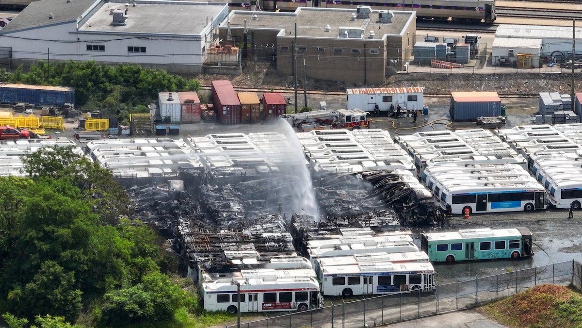 An aerial view of a large parking lot at SEPTA's Midvale Depot in North Philadelphia. There are dozens of decommissioned SEPTA buses tightly and neatly packed in rows, bumper to bumper and side to side. A group of them had clearly been on fire, and are being sprayed from afar with water from a fire hose and fire truck. They are very badly burned, and they look like blackened shells of buses.