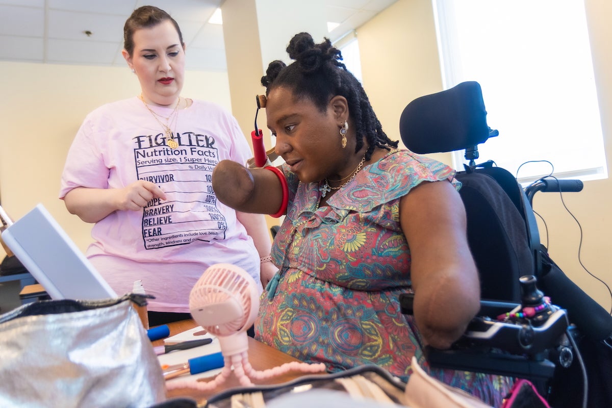 During a support group session for people who had traumatic health events at Philadelphia's Jefferson rehabilitation center, Candice Davis, who is recovering after having all four limbs amputated do to complications from COVID-19, is sitting in a wheelchair, watching herself closely in a mirror as she attempts to put on makeup using a device attached the small portion of her right arm that remains. A nurse is standing next to her, offering advice and guidance.