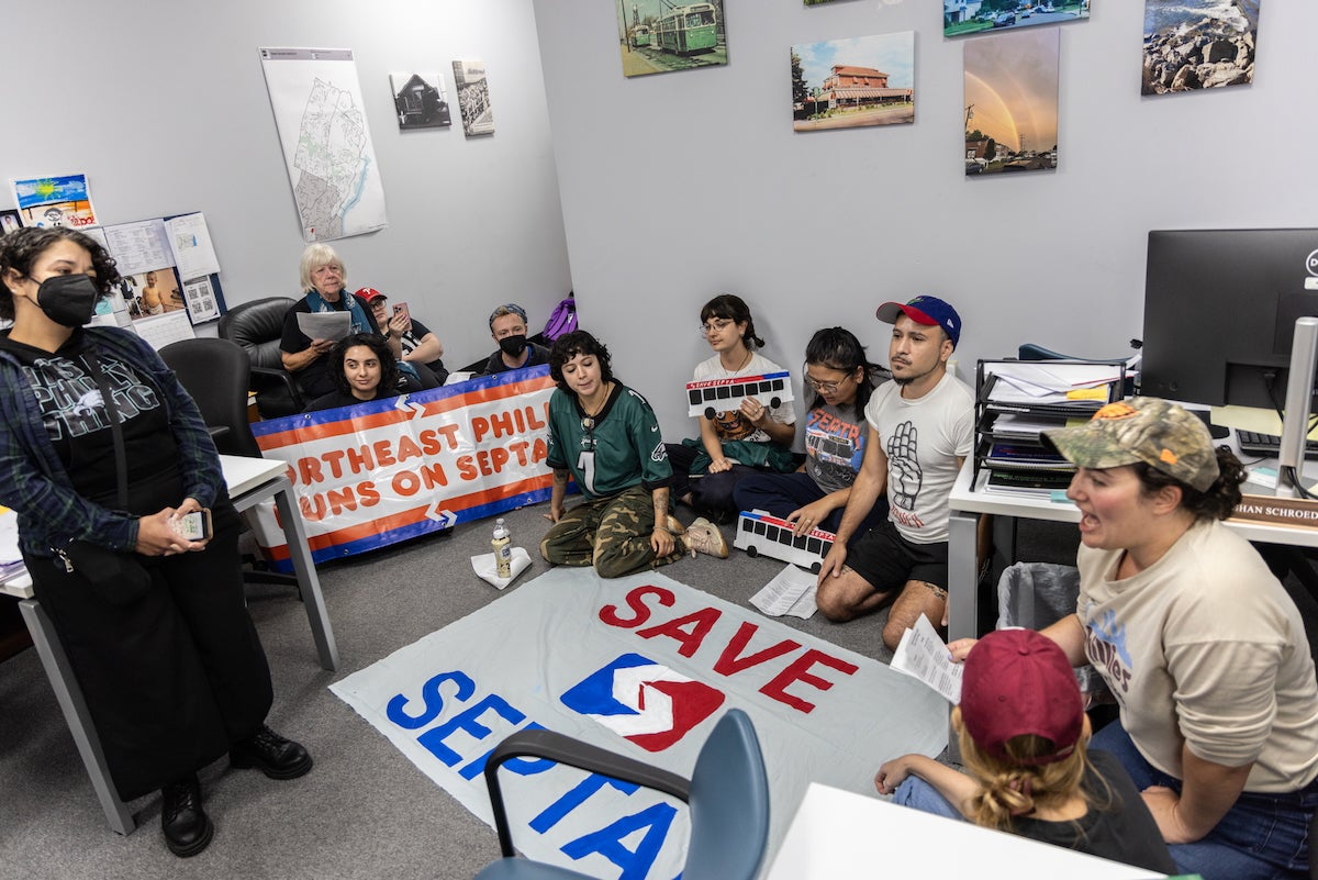 A group of 9 protesters from 215 People Alliance is staging a sit-in protest in the office of Republican Pa. Senator Joe Picozzi in Philadelphia. A large sign is on the floor, which says 'Save SEPTA', and another person holds a sign that says 'NORTHEAST PHILLY RUNS ON SEPTA.