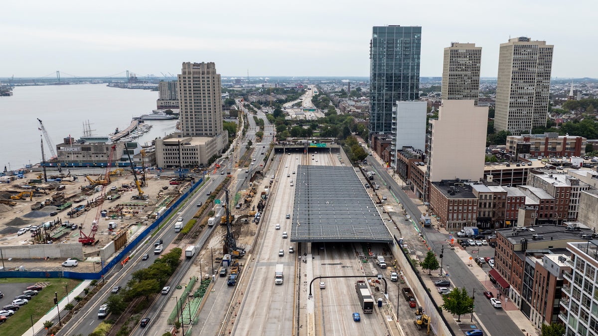 An aerial view of I-95 in Philadelphia, with the Delaware River to the left. Construction is happening to build a cap over the highway, and so far, one side of the highway is completely covered for a small stretch.