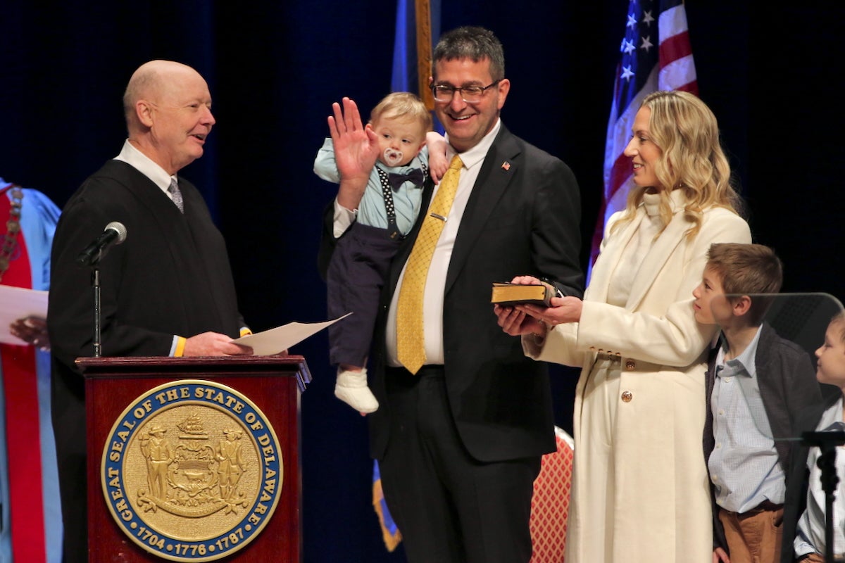 Standing behind a podium with the state seal of Delaware on the front at Delaware State University, State Supreme Court Justice Collins J. Seitz, Jr., is swearing in Gov. Matt Meyer. Meyer has his right hand raised while also holding onto an infant. Next to him are his wife and two other children.