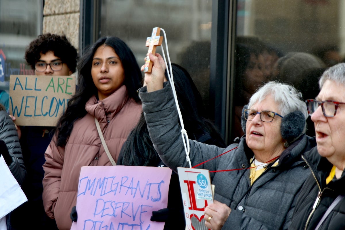 Outside Philadelphia's ICE office in Chinatown, a group of protesters stands side by side. Two of them hold up signs reading 'Immigrants deserve dignity,' and one woman holds up a cross.