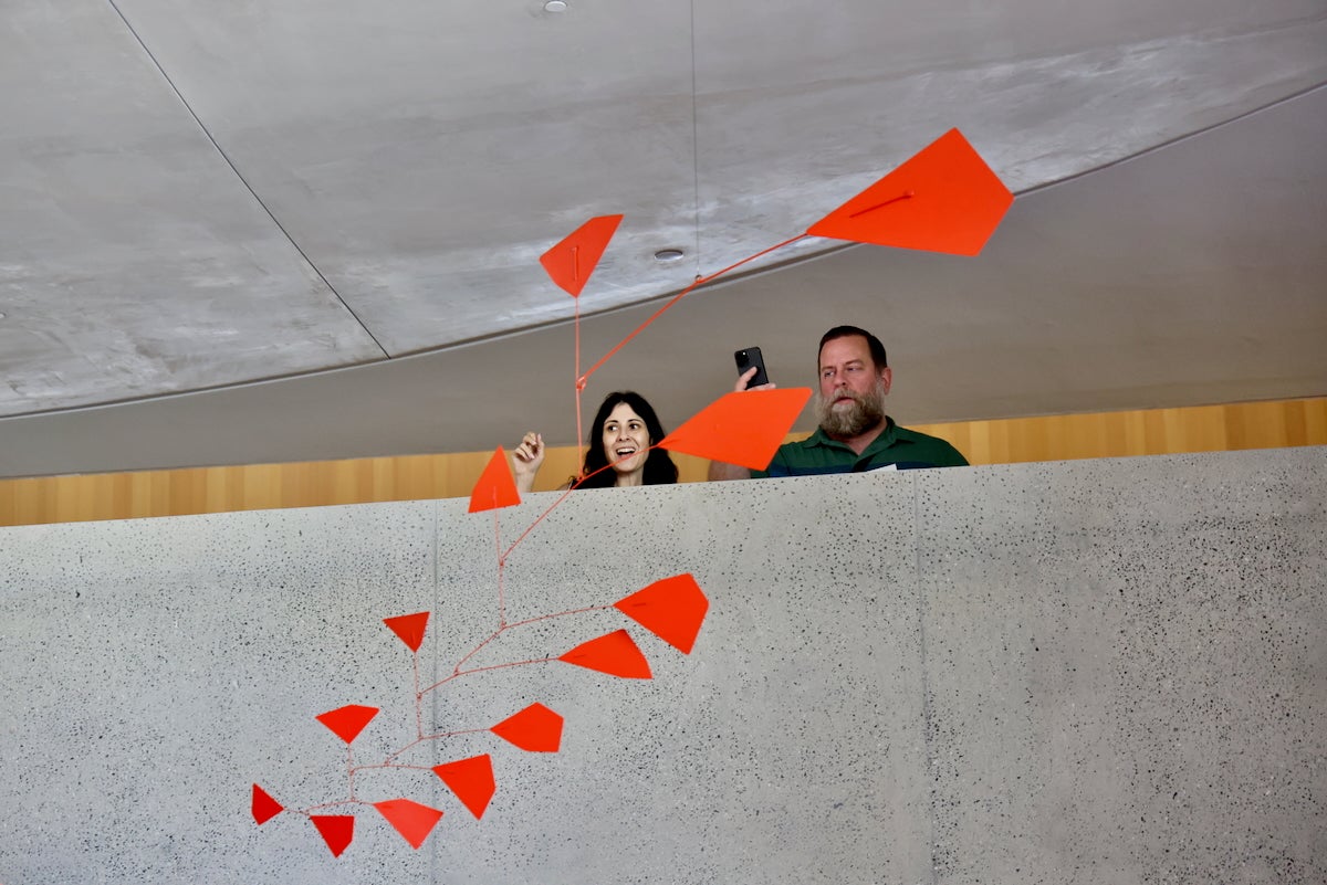 Two people are on a balcony, looking down at a constantly shifting hanging red sculpture made by Alexander Calder at the Open Plan Gallery in the newly opened Calder Gardens in Philadelphia.