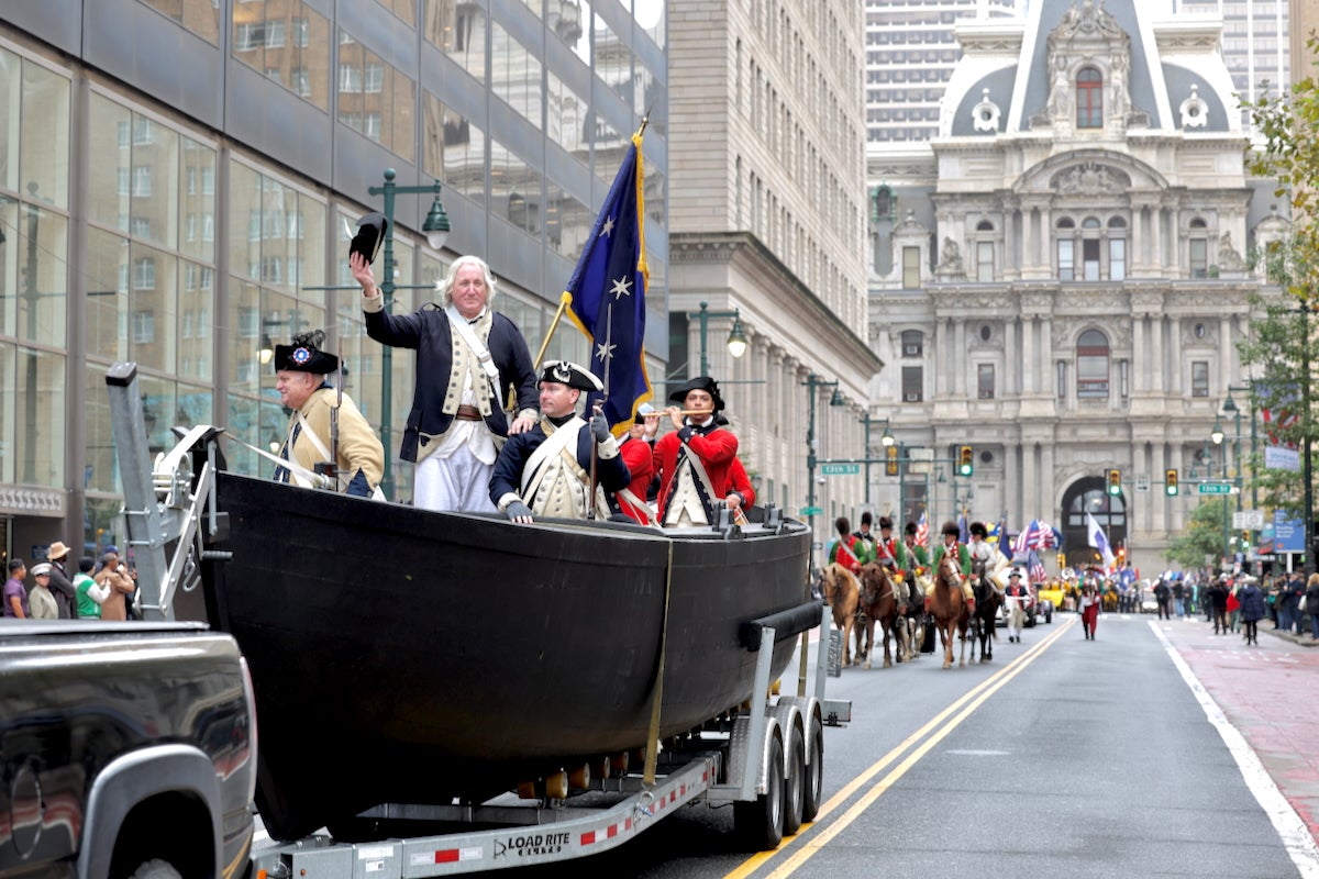 Historical reenactors dressed up as George Washington and his troops are in a replica of the Durham boats that carried Washington across the Delaware to victory at Trenton in 1776. They are being pulled by a van as they travel on the street away from Philadelphia City Hall.