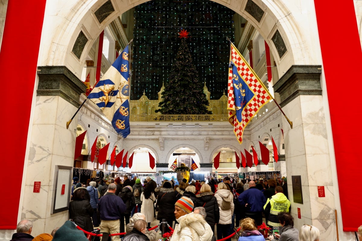 Many people are crowded in the Grand Court at the Wanamaker Building in Center City Philadelphia, watching the traditional Christmas light show, which is occurring on the wall of the very high-ceilinged space.