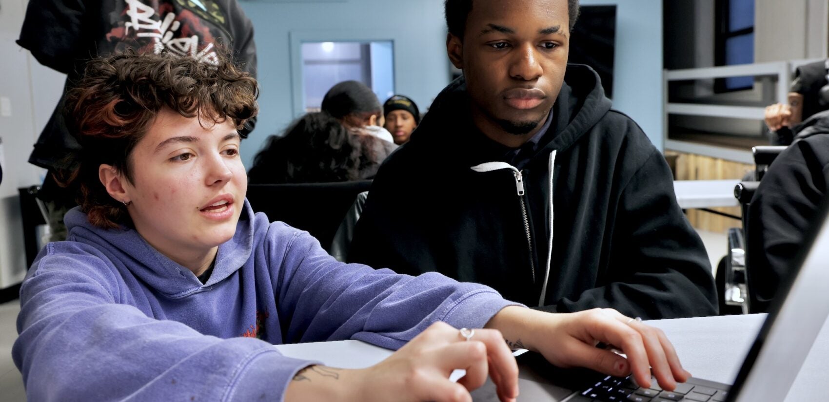 Noelle Correa sitting with Isaiah Blackwell, watching as they are working on a computer in the classroom