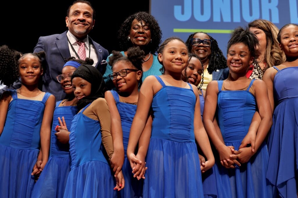 A group of young girls from the Edward Gideon School Junior Dance Ensemble, wearing matching blue dresses, is on stage with Cherelle Parker after performing a dance routine.