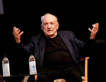 Architect Frank Gehry sits on a stage with his hands extended outward and upward
