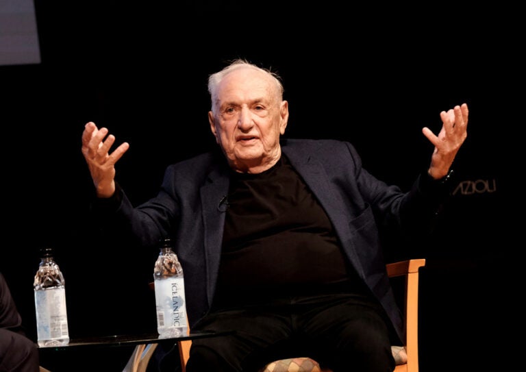 Architect Frank Gehry sits on a stage with his hands extended outward and upward